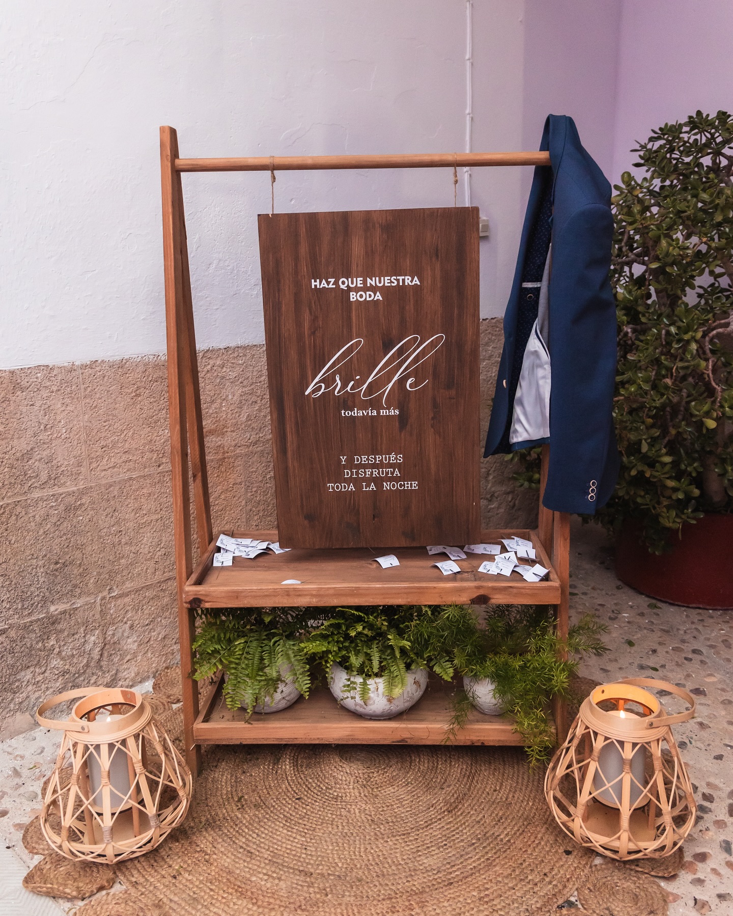 Cartel de bienvenida a boda con mensaje personalizado, acompañado de plantas y farolillos decorativos sobre alfombra de yute.