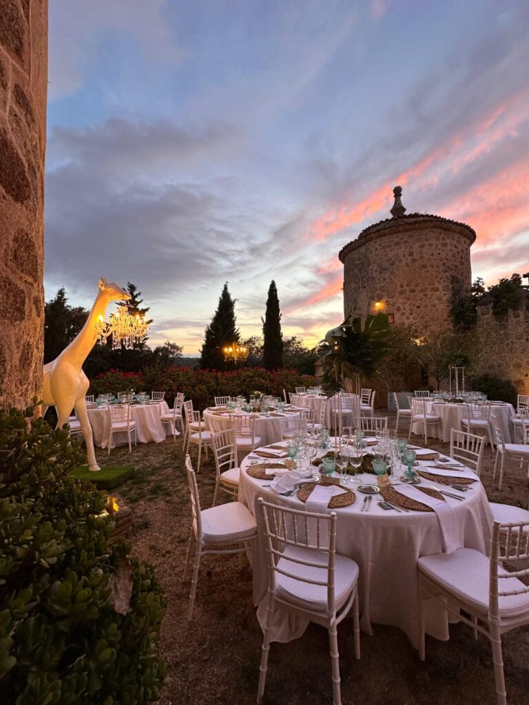 Montaje de boda al atardecer en jardín con mesas redondas, sillas blancas y lámparas colgantes, con cielo en tonos cálidos.